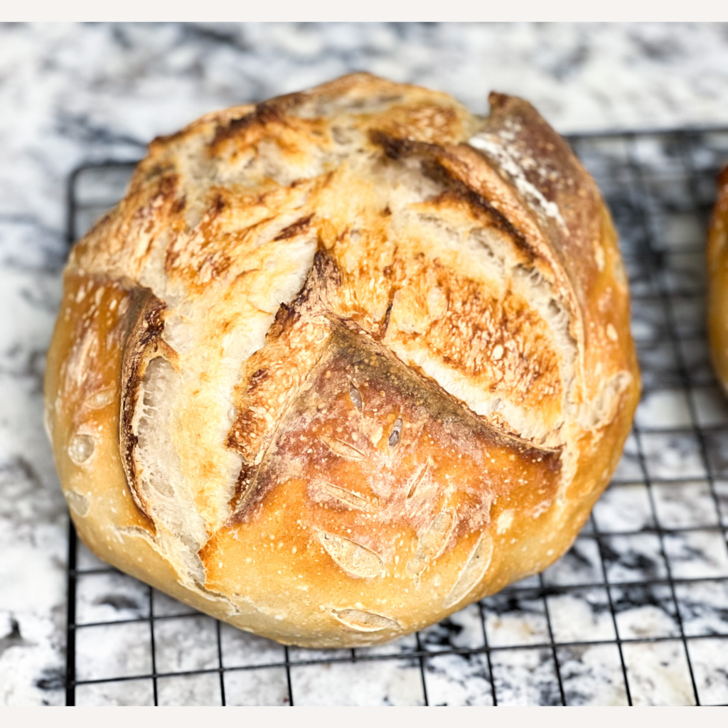 Crusty round sourdough boule with decorative scoring, cooling on a wire rack over a marble countertop.